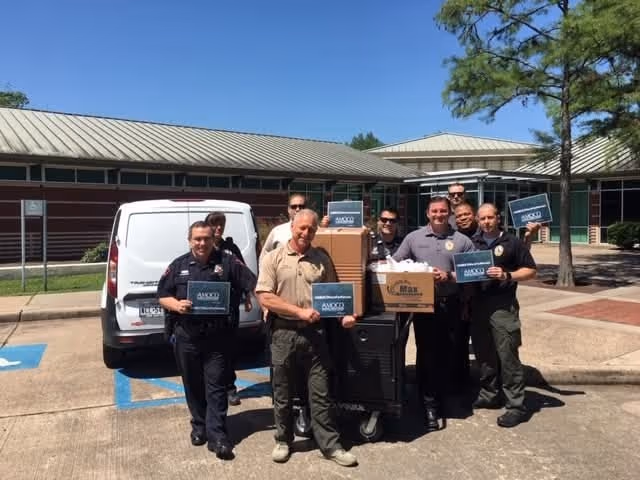A group of law enforcement officers stands outside a building, holding boxes and signs in front of a parked van on a sunny day.