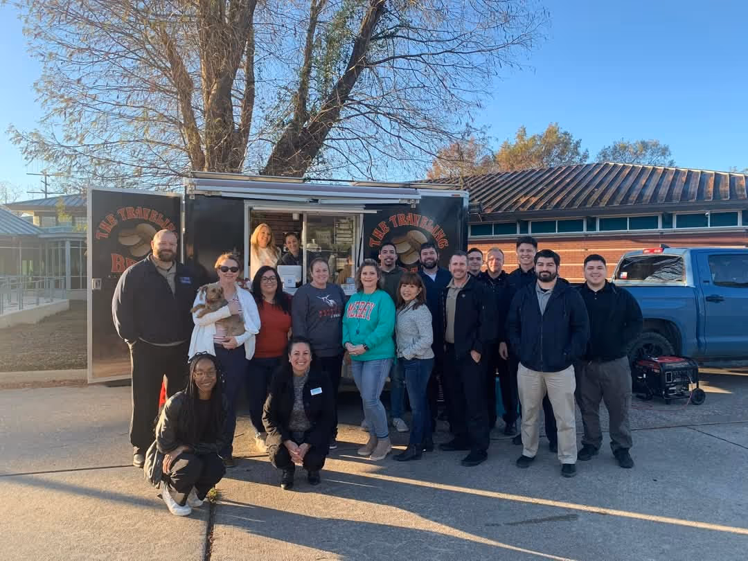 A diverse group of people gathered in front of a food truck, smiling, with a tree and a pickup truck in the background.