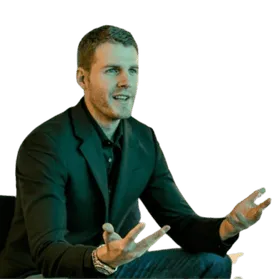 A man in a black blazer passionately gestures while speaking, seated against a neutral background.
