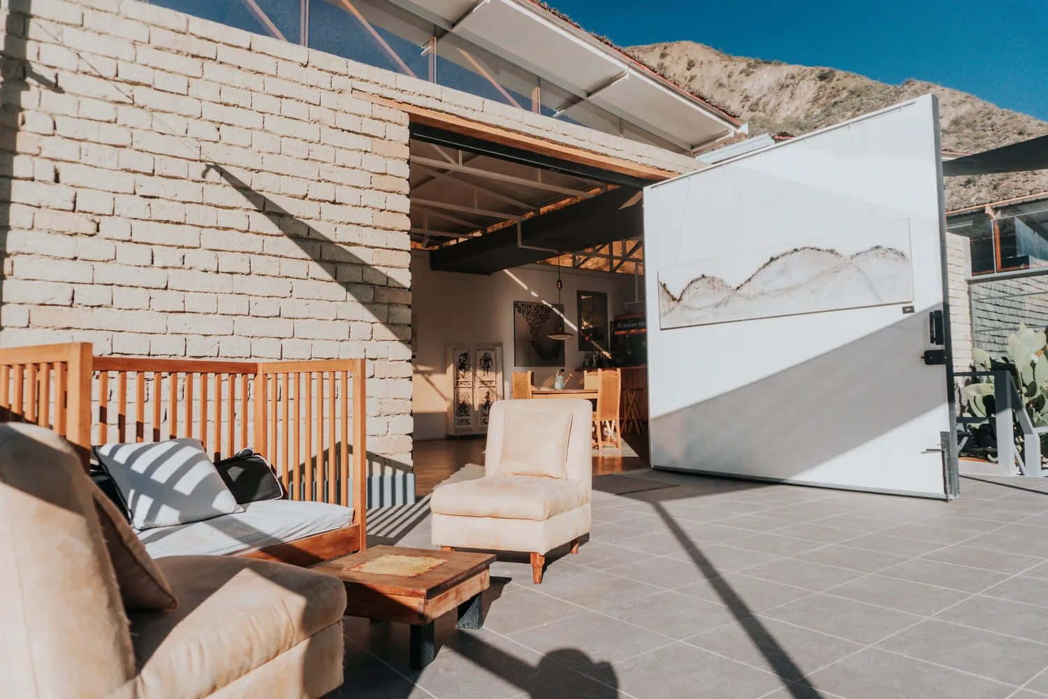 Terraza con sillón y sofá de madera frente a una puerta abierta, junto a un muro de ladrillo.