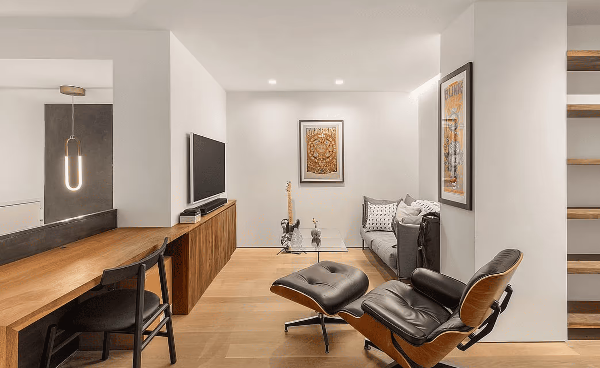 Modern living room with black leather lounge chair, gray sofa, glass coffee table, electric guitar, and wooden desk with chair under hanging light.