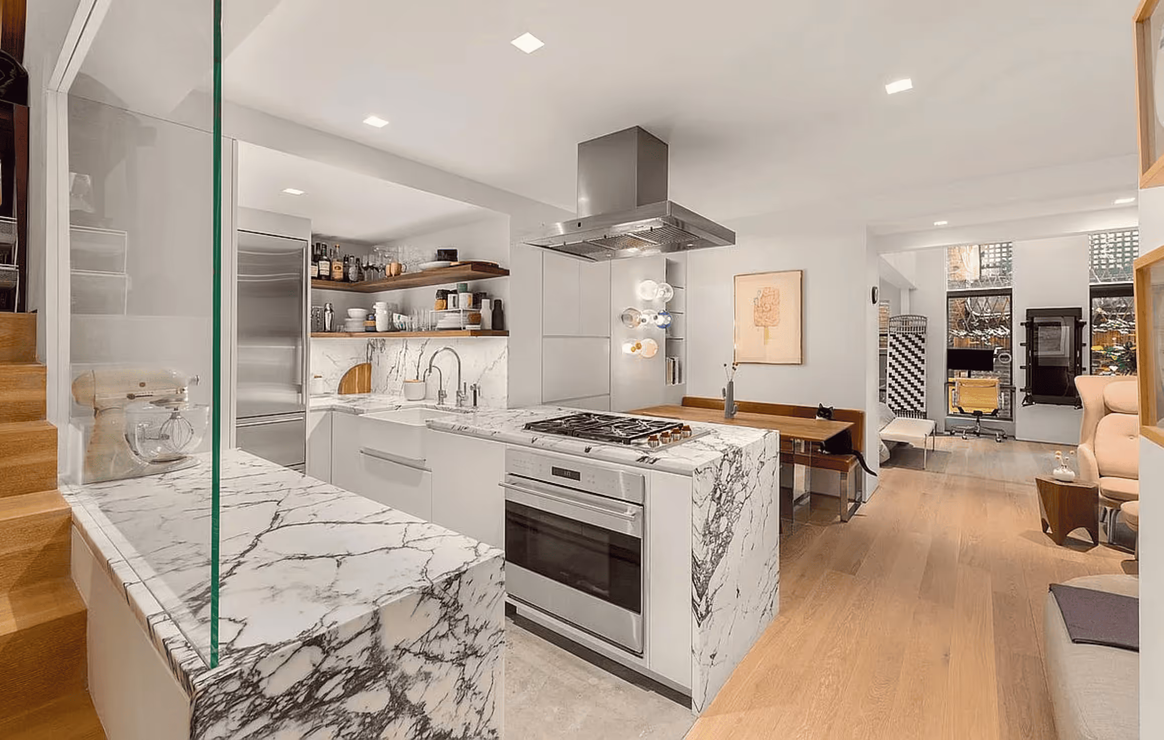 Modern kitchen with marble countertops, stainless steel appliances, wooden shelves, and an open living area with wooden flooring.