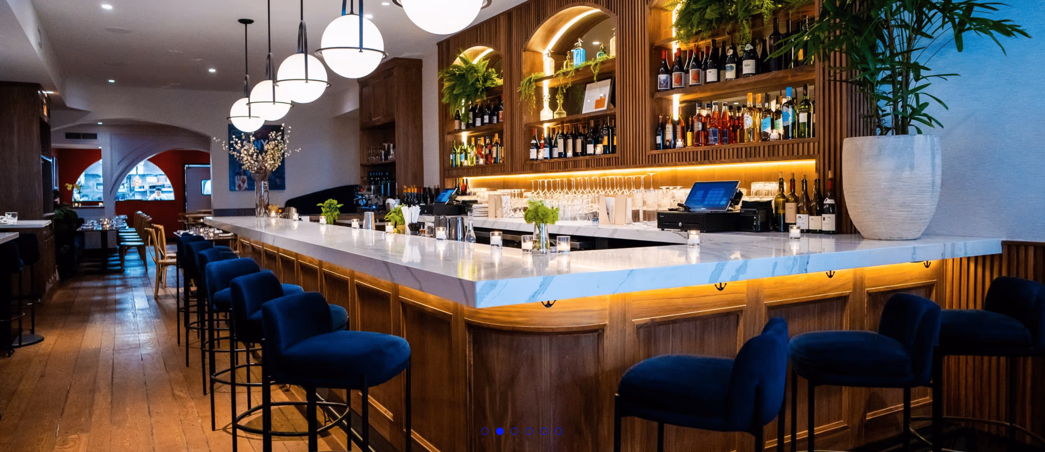 Modern bar with a white marble countertop, blue velvet bar stools, wood flooring, and shelves stocked with bottles and glassware.