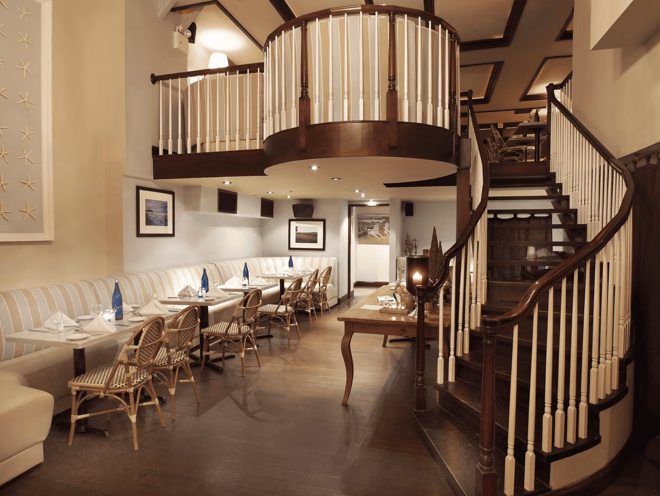 Cozy restaurant interior with a curved wooden staircase, white banquette seating, and tables set with napkins and water bottles.