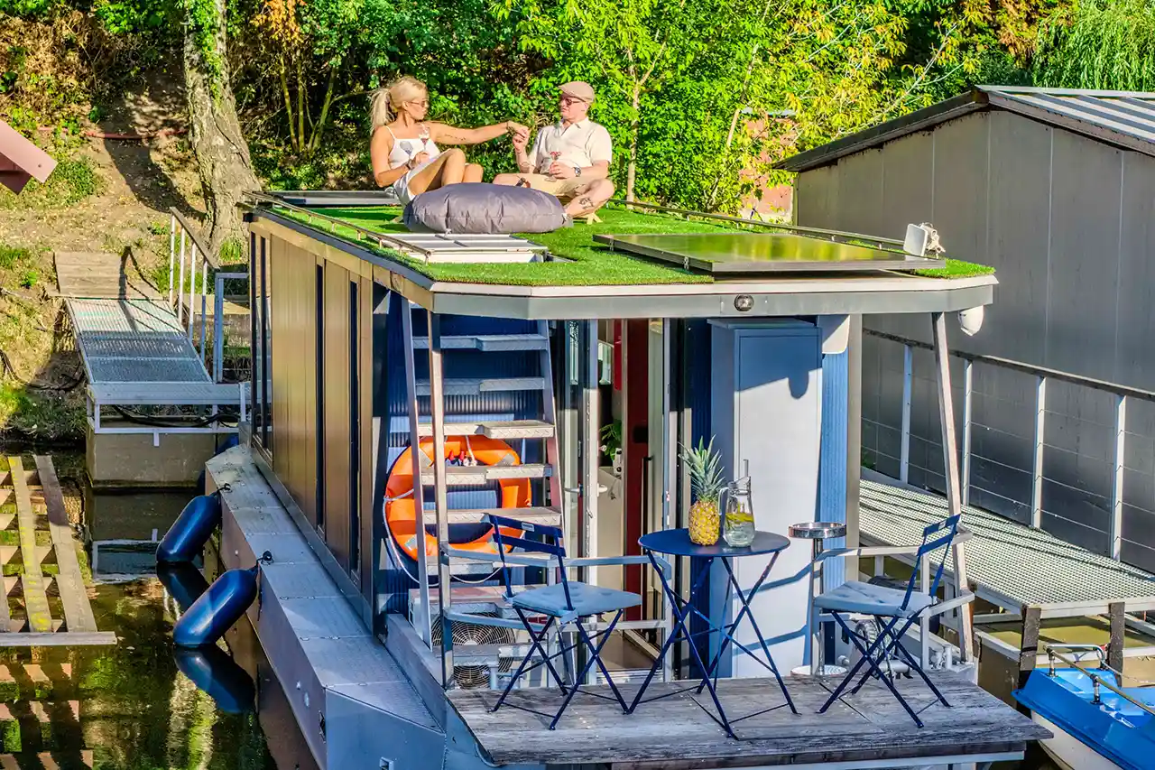 Pár si užívá snídani na zelené střešní terase houseboatu Neptun s výhledem na Vltavu. Dole paluba se stolkem, židlemi a záchranným kruhem.