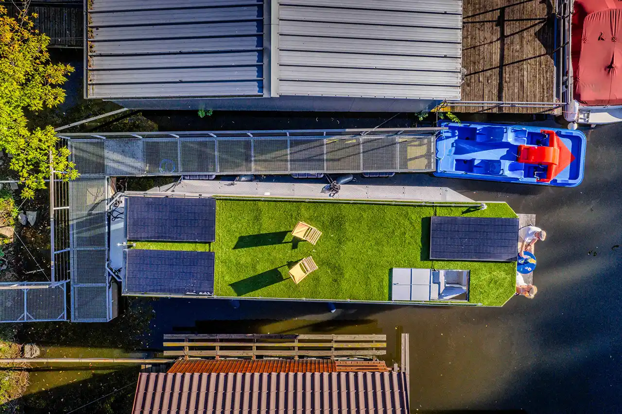 Pár si užívá snídani na zelené střešní terase houseboatu Neptun s výhledem na Vltavu. Dole paluba se stolkem, židlemi a záchranným kruhem.