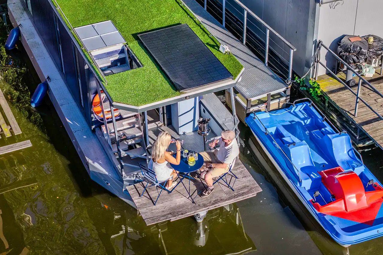 Pár připíjí vínem na palubě houseboatu Neptun při západu slunce. Vedle kotvící modré šlapadlo, v pozadí schody na střešní terasu.