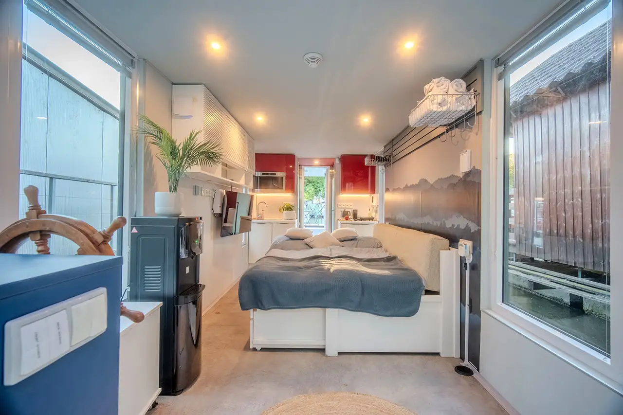 Interiér houseboatu Neptun – manželská postel s výhledem na řeku, kuchyňský kout, lodní kormidlo a prostorné okna po obou stranách.