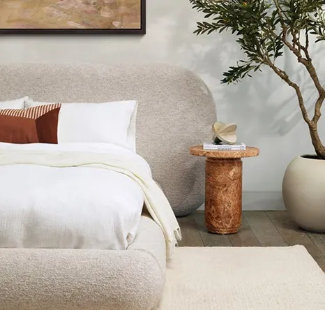 White mattress with white pillows and grey puffy headboard, with marble brown side table next to tall house plant in a large grey pot. 