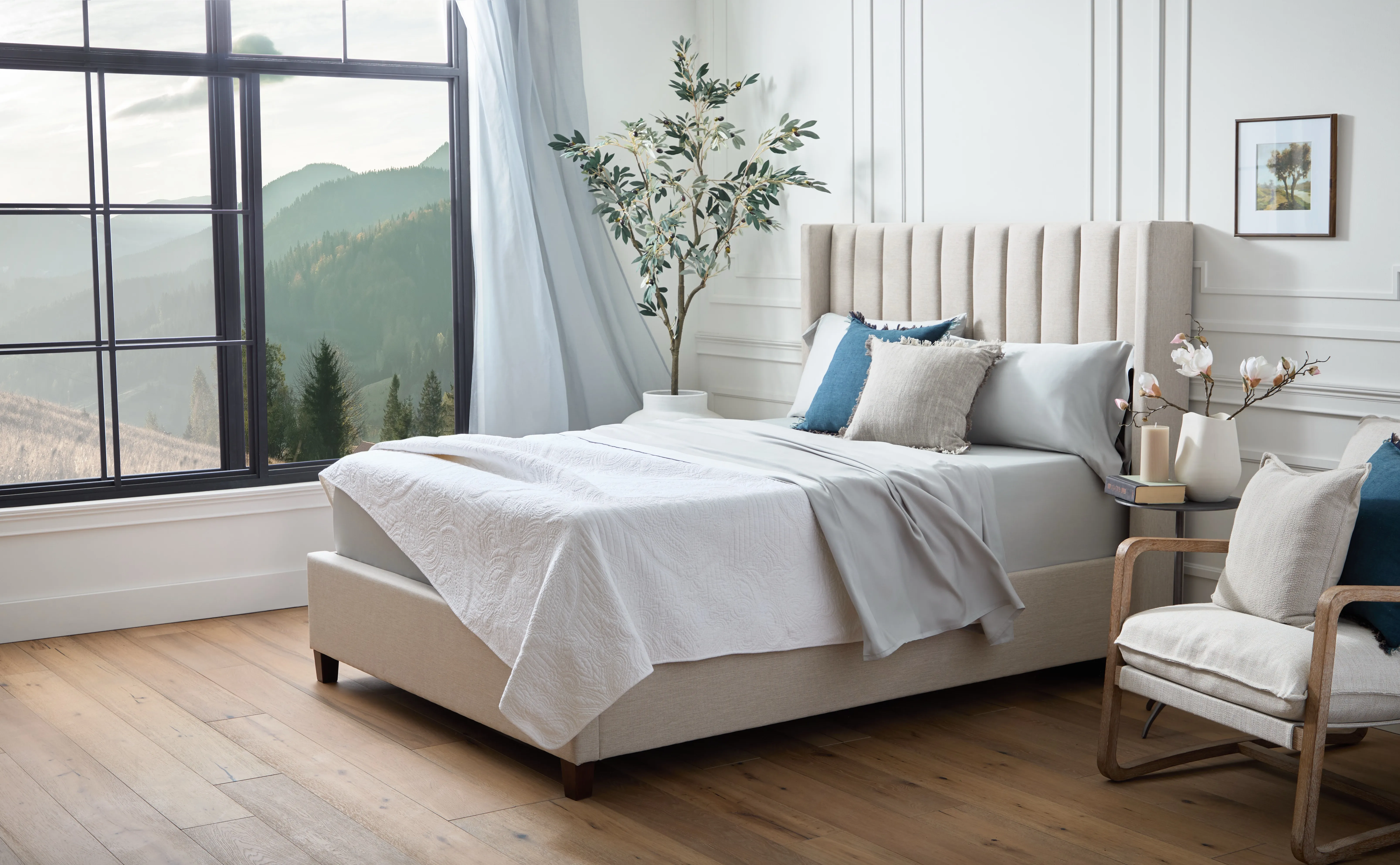 Brightly lit bedroom with green mountains in window, light grey tall headboard, white comforter, and tall plant in the corner next to bed