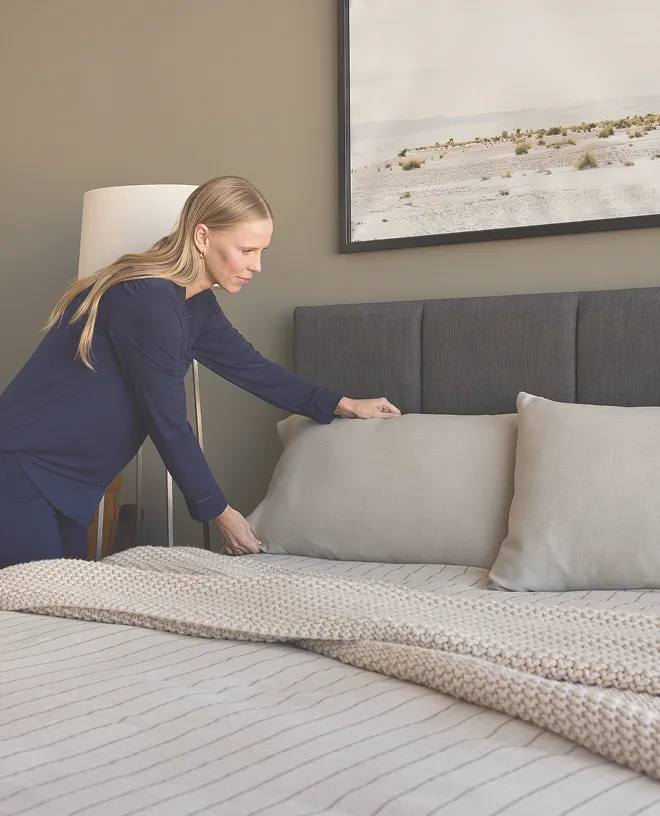 Blonde woman with long hair in blue long sleeves adjusting grey pillow on bed