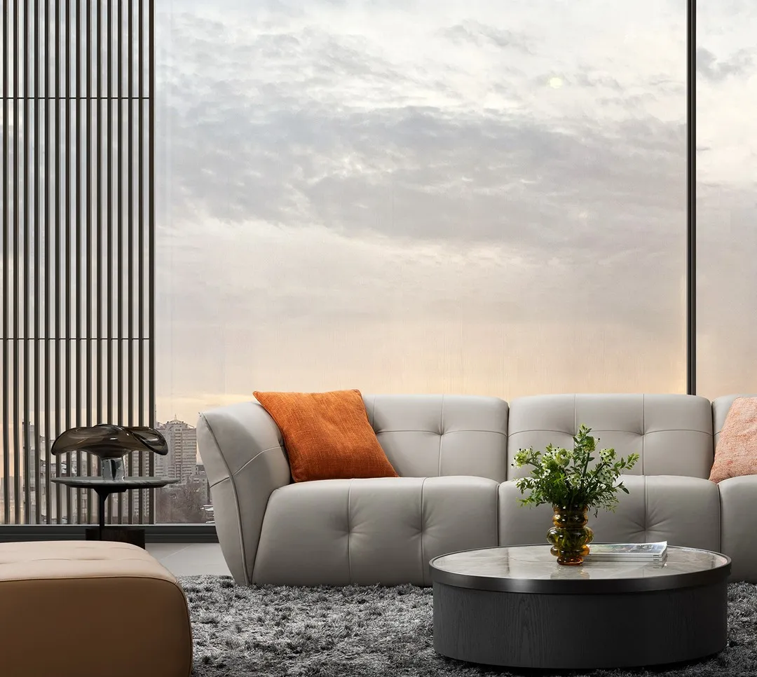 Light grey sofa with one orange pillow with round coffee table and large window in the background 