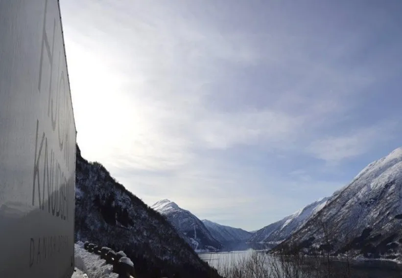 Blue sky and image of fjords, snowy mountains, and lake that reflects the sun. 