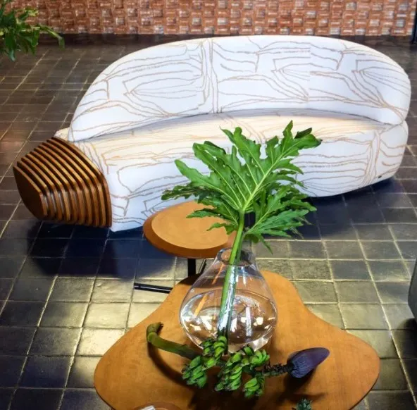 Modern curved sofa with abstract-patterned white upholstery and a sculptural wooden accent on one end, placed on a dark tiled floor with a mid-century style coffee table and tropical plant in the foreground