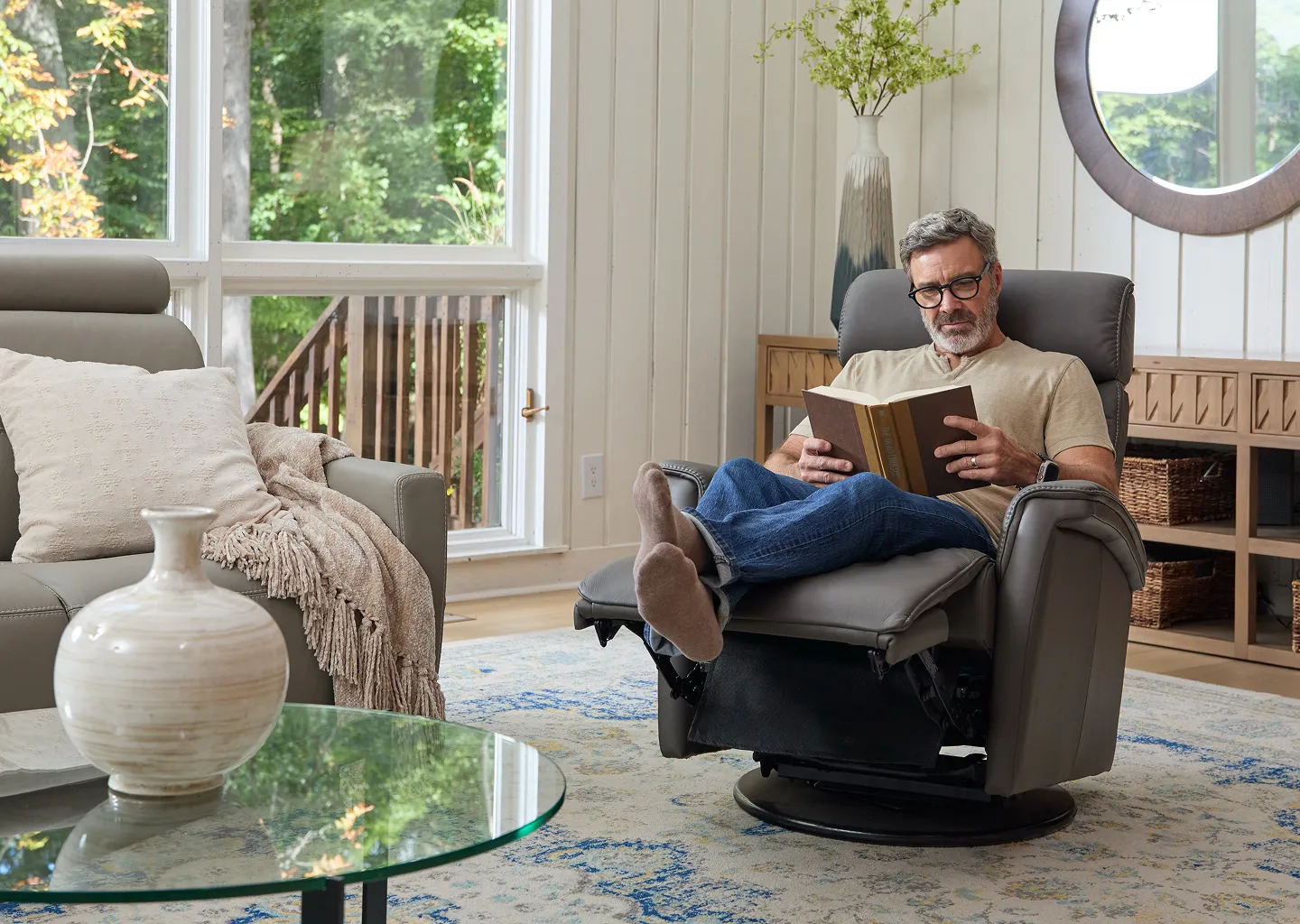 Man with grey hair and black glasses intently reading a book sitting in a grey recliner with his feet up in jeans and a light yellow t-shirt 