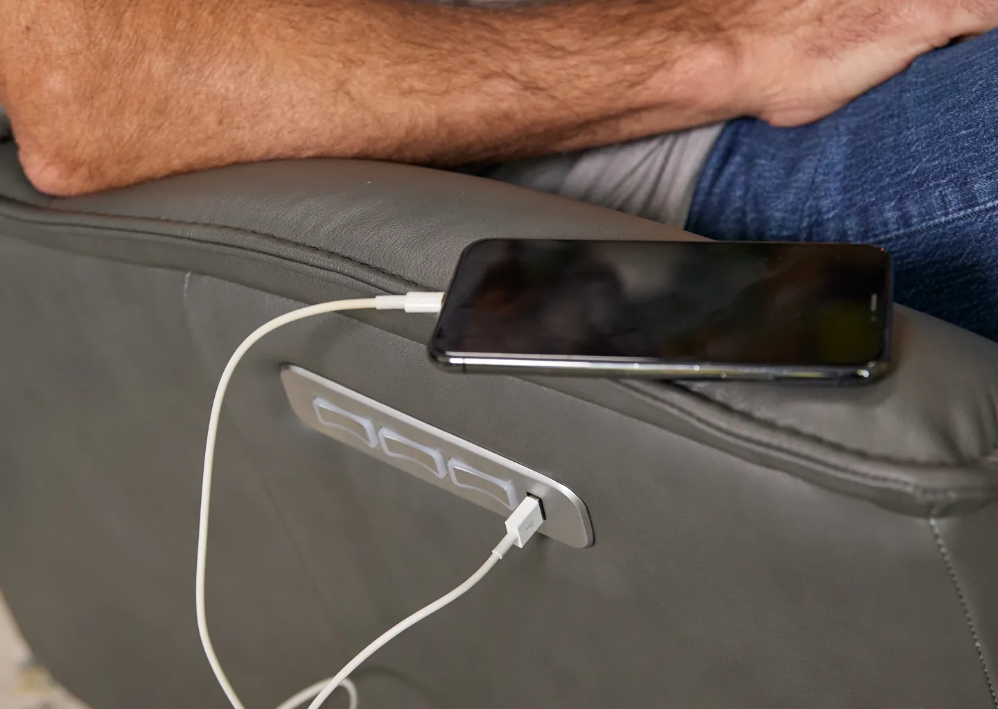 iPhone sitting on top of dark grey leather recliner arm, iPhone is plugged into built-in charger in the side of recliner