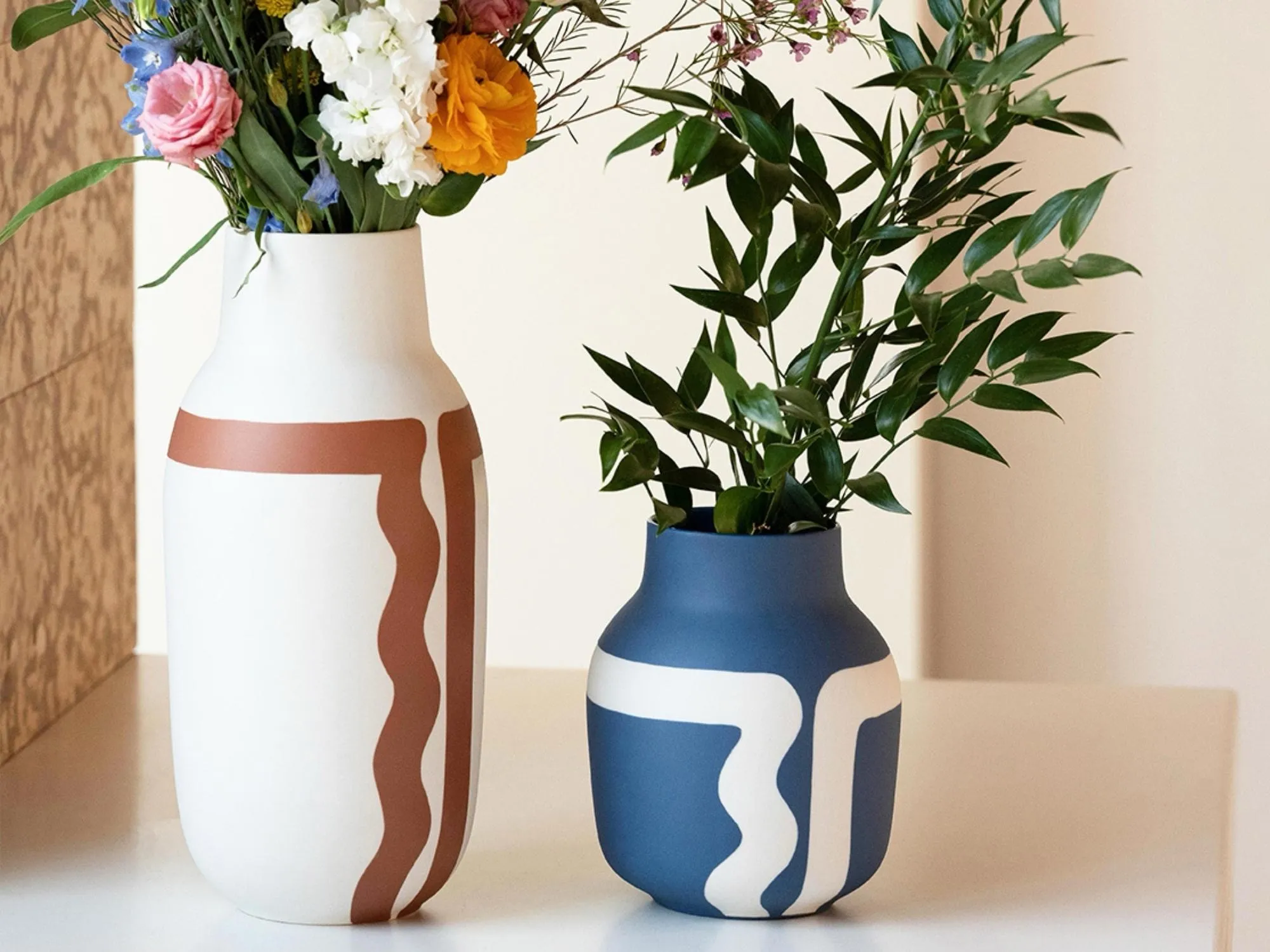 Two vases sitting on a shelf, one white with a brown abstract design, the other blue with a white abstract design - wax painted by hand and feature a glazed interior. Both holding flowers.