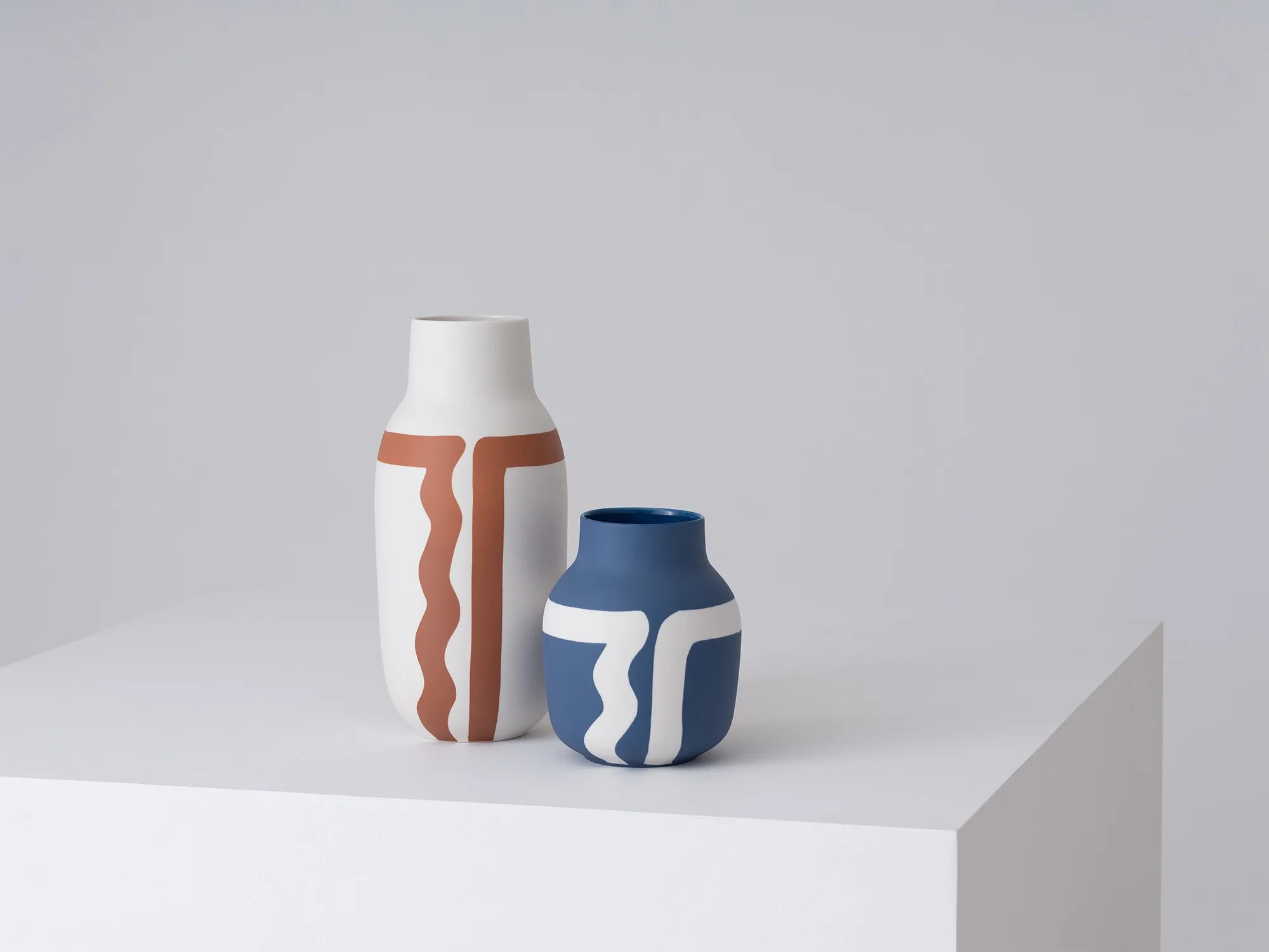 Two hand painted vases with a white background, the large white vases has a abstract brown pattern, the blue smaller vase has a abstract white pattern. 