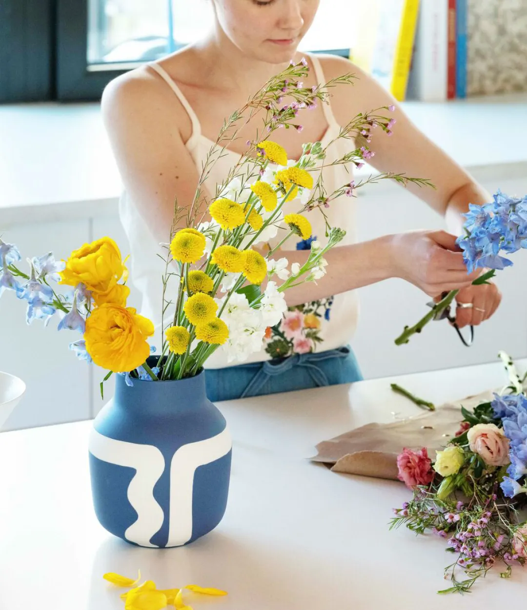 A woman cutting blue flowers to put into a blue and white vase with tall yellow flowers sitting in the vase 