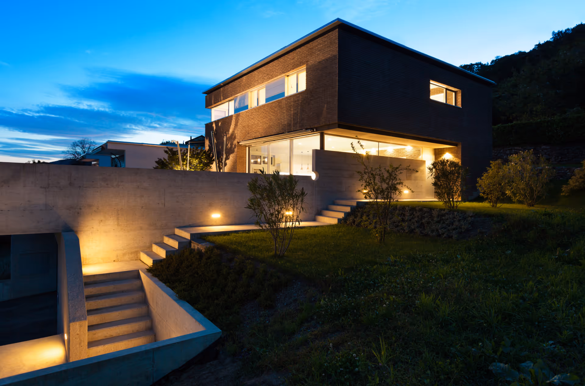 Early evening shot of house built on to hill with steps and lit driveway. 