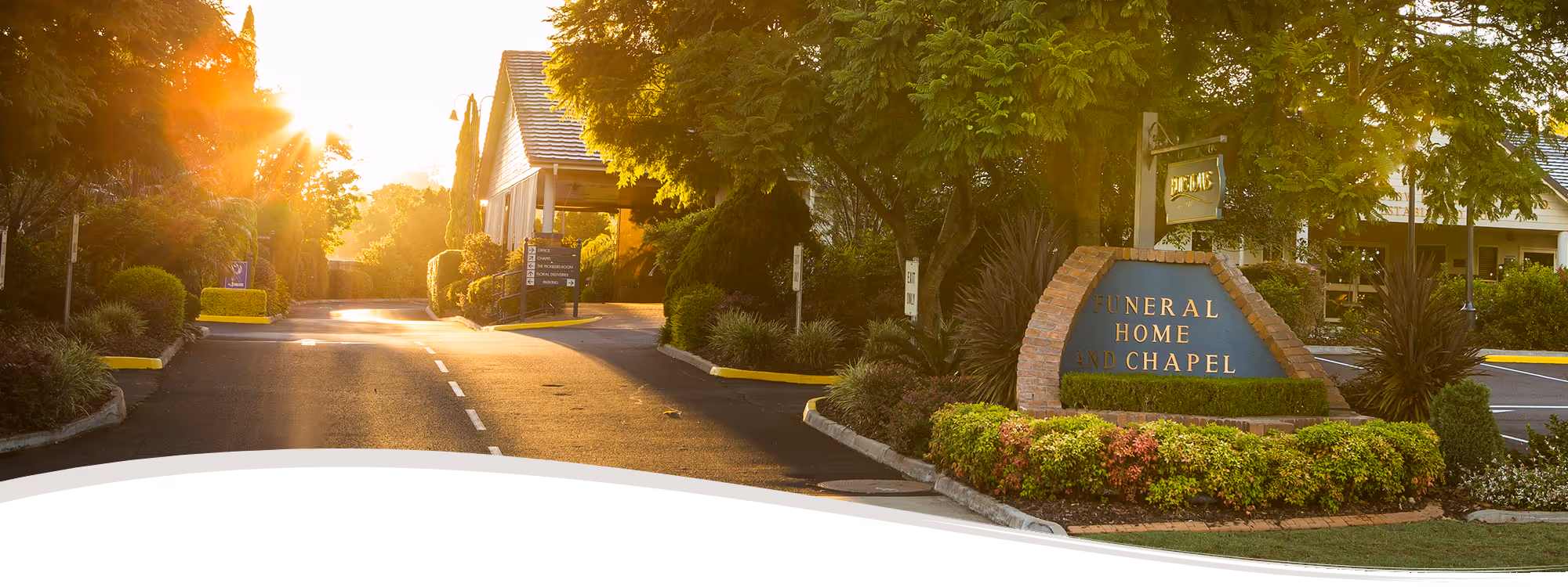 A long-distance view of a funeral home and chapel is shown at sunset. The entrance is marked by a large, stone sign on the right side of the road with the words "Funeral Home & Chapel" etched into it. The sun is setting behind the trees and buildings on the left, casting a warm glow and long shadows on the paved driveway. The buildings have a rustic, homey feel, with a visible roof and some windows. In the background, there are trees and shrubs on either side of the road.