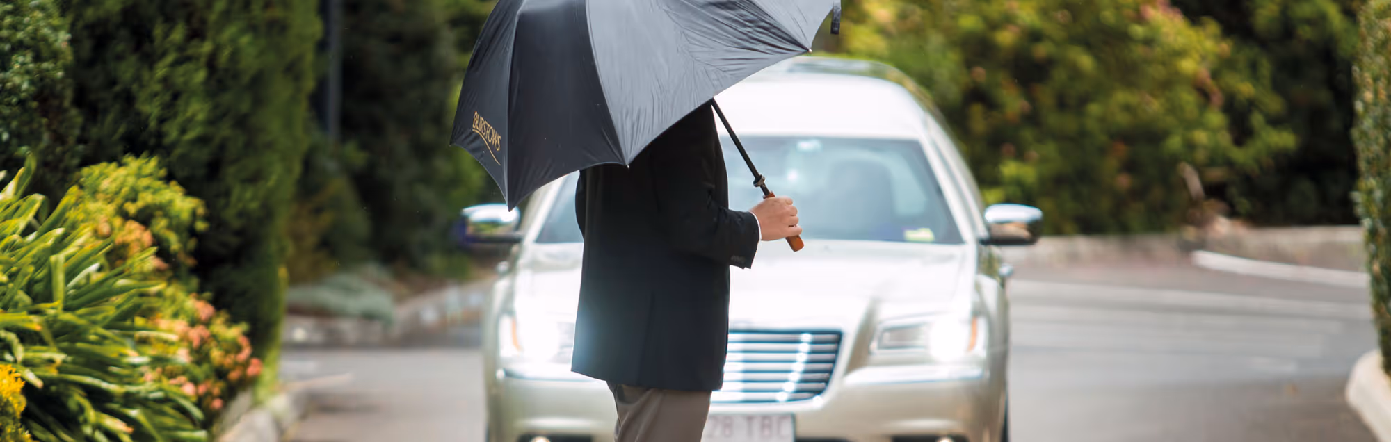 A funeral attendant holds an umbrella by a hearse on a rainy day.