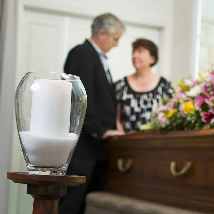 A memorial candle with a blurred couple and casket in the background.