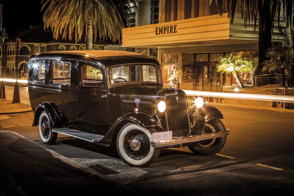 A vintage black car with white-wall tires parked at night in front of a building with an "EMPIRE" sign.