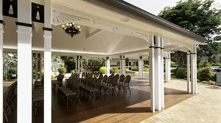 A covered, outdoor area with rows of chairs is the focus of this image. The structure is supported by white and black columns, with intricate trim and an elegant chandelier hanging from the ceiling. The floor is made of wood, and the chairs are a dark brown color. In the background, there is a manicured lawn, shrubs, and a building with a large window. This looks like an outdoor memorial or funeral service venue.