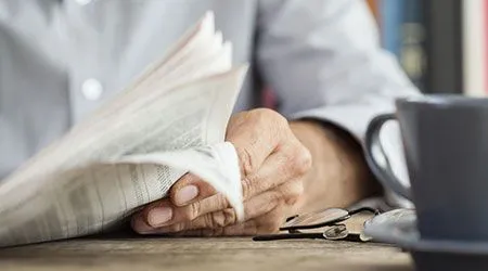 
A person's hands are shown holding a folded newspaper, with their left hand holding the pages open while their right hand is resting on a wooden table. Next to their right hand, there are a pair of glasses and a small pile of coins. To the right of the coins, a gray mug sits on a saucer.