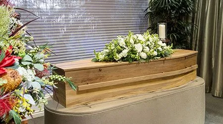 A small, light wooden casket rests on a soft, cushioned stand. A white floral arrangement, including roses and other flowers, is placed on top. A single white candle in a glass holder is visible behind the casket. To the left, there is another large bouquet of flowers with red and green blooms. The setting appears to be a room within a funeral home or a chapel.
