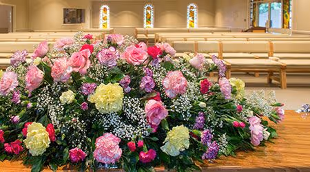 
A large floral arrangement rests on what appears to be a casket, though the casket itself is not visible. The arrangement is made up of a mix of pink and yellow flowers, likely carnations and roses, with smaller white flowers mixed in.

In the background, there is a large room with several rows of wooden pews and three tall, arched stained-glass windows. This setting suggests the image was taken in a church or chapel, likely during a funeral service.