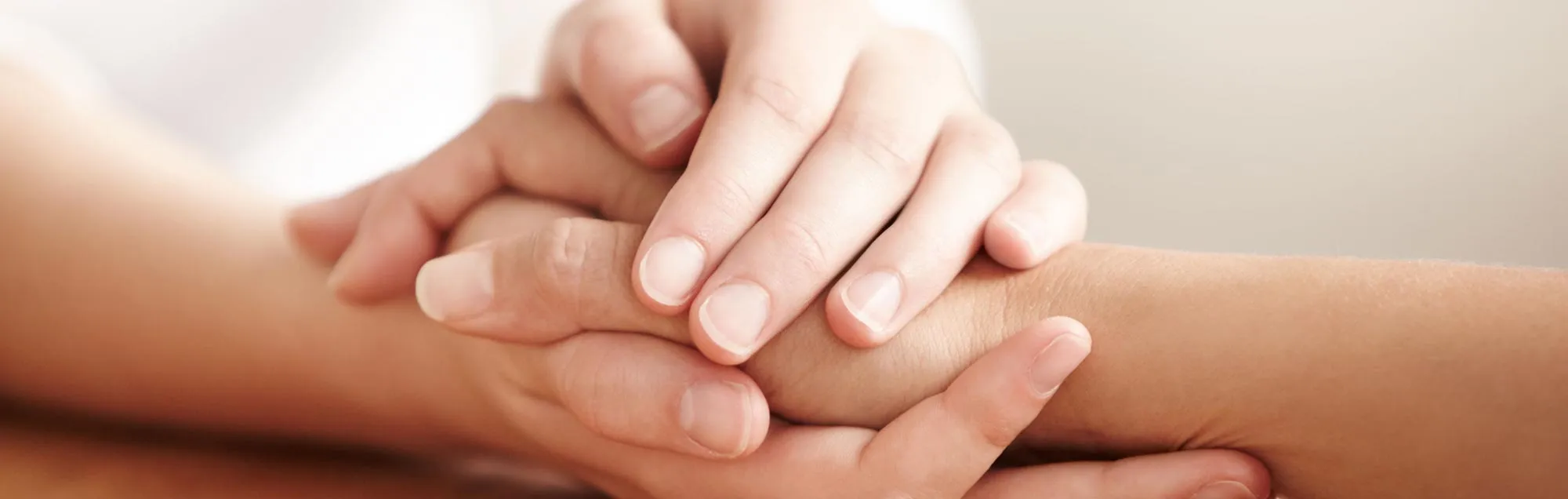 
A close-up image shows three hands clasped together, creating a sense of comfort and support. The top hand is gently resting on the forearm and hand of another person below. The fingers are intertwined, showing a connection between the individuals. The image is cropped to focus on the hands, with a warm, soft background. The light tone of the image suggests empathy and solace.