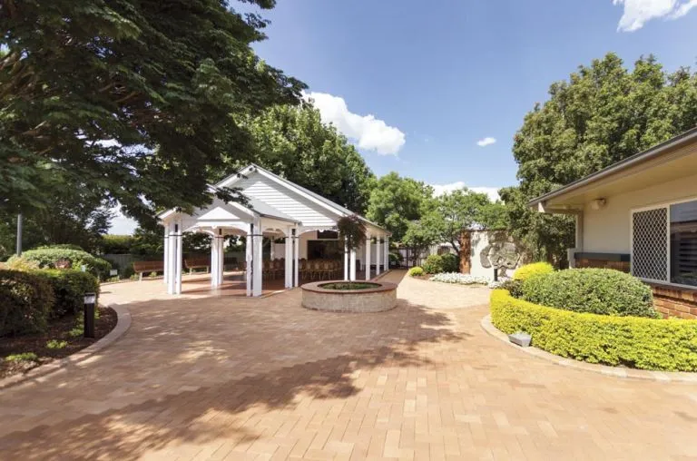 A sunny outdoor space features a covered gazebo with a white roof and columns, and a brick-paved courtyard. The gazebo has chairs and tables set up inside, with benches visible on the left. In the center of the courtyard, there is a circular brick planter. The surrounding area is landscaped with manicured hedges, shrubs, and large trees. On the right, a building is visible with large windows. The overall setting appears to be a formal outdoor space for events or gatherings, possibly at a funeral home.