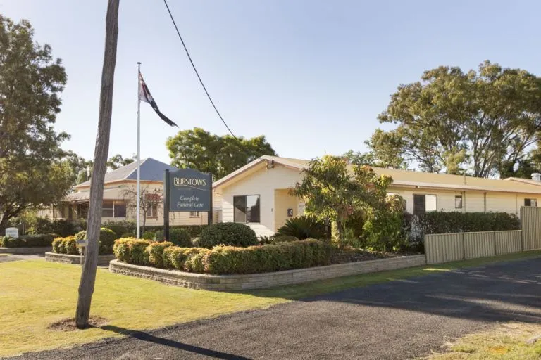 
A low-rise, beige building with a sloped roof is shown on a sunny day. A sign in front of the building reads "BURSTOWS Complete Funeral Care." There is a flagpole with a flag flying on the left side of the image. The building is surrounded by a well-manicured lawn and hedges, and a paved driveway leads up to the entrance. This looks like a typical funeral home with a private, residential feel.