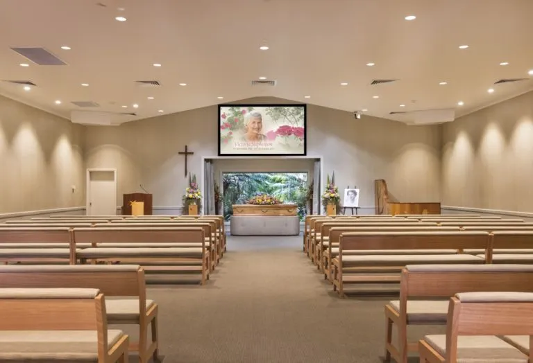 A modern funeral chapel or crematorium is shown from the back of the room, looking toward the front. The room is clean and well-lit, with rows of wooden pews on a carpeted floor. At the front, there is a large screen showing a photo of a woman with a floral border. Below the screen is a casket with a floral arrangement on top. To the right, an easel holds a portrait, and a piano sits nearby. A wooden pulpit and a cross hang on the wall to the left.