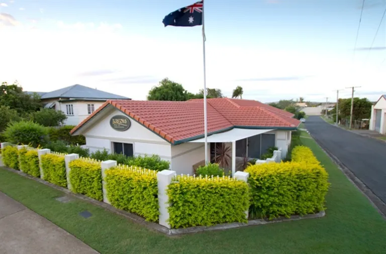 
A one-story building with a red-tiled roof and a flagpole is shown from an elevated perspective. The building is surrounded by a well-trimmed hedge and a white fence. A sign is visible on the building's facade with the name "BURSTOWS." An Australian flag is flying from the flagpole. The setting appears to be a quiet residential neighborhood, with a street on the right side of the image. This is a funeral home in what looks like a converted house.