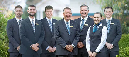 A group of seven people, six men and one woman, are standing together and smiling at the camera. They are all formally dressed in dark suits, white shirts, and ties, with the woman wearing a vest and scarf. Each person is wearing a name tag on their lapel. The setting appears to be outdoors in front of a building and some greenery.