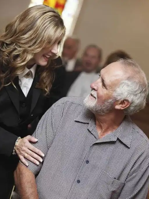 A funeral director in a black suit comforts a seated elderly man during a memorial service.
