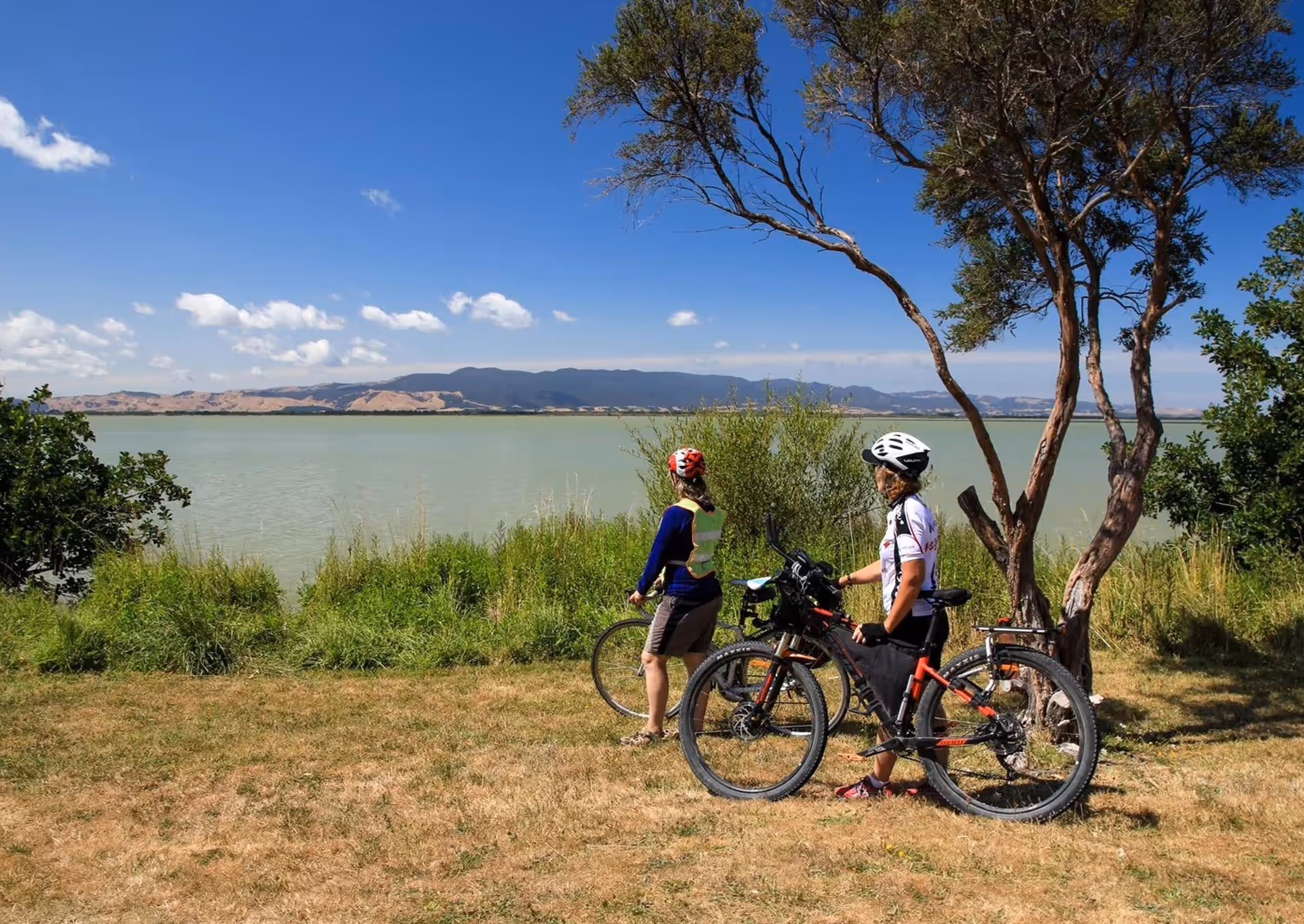 Two cyclists wearing helmets standing with their bikes by a lakeside with mountains in the background under a clear blue sky.