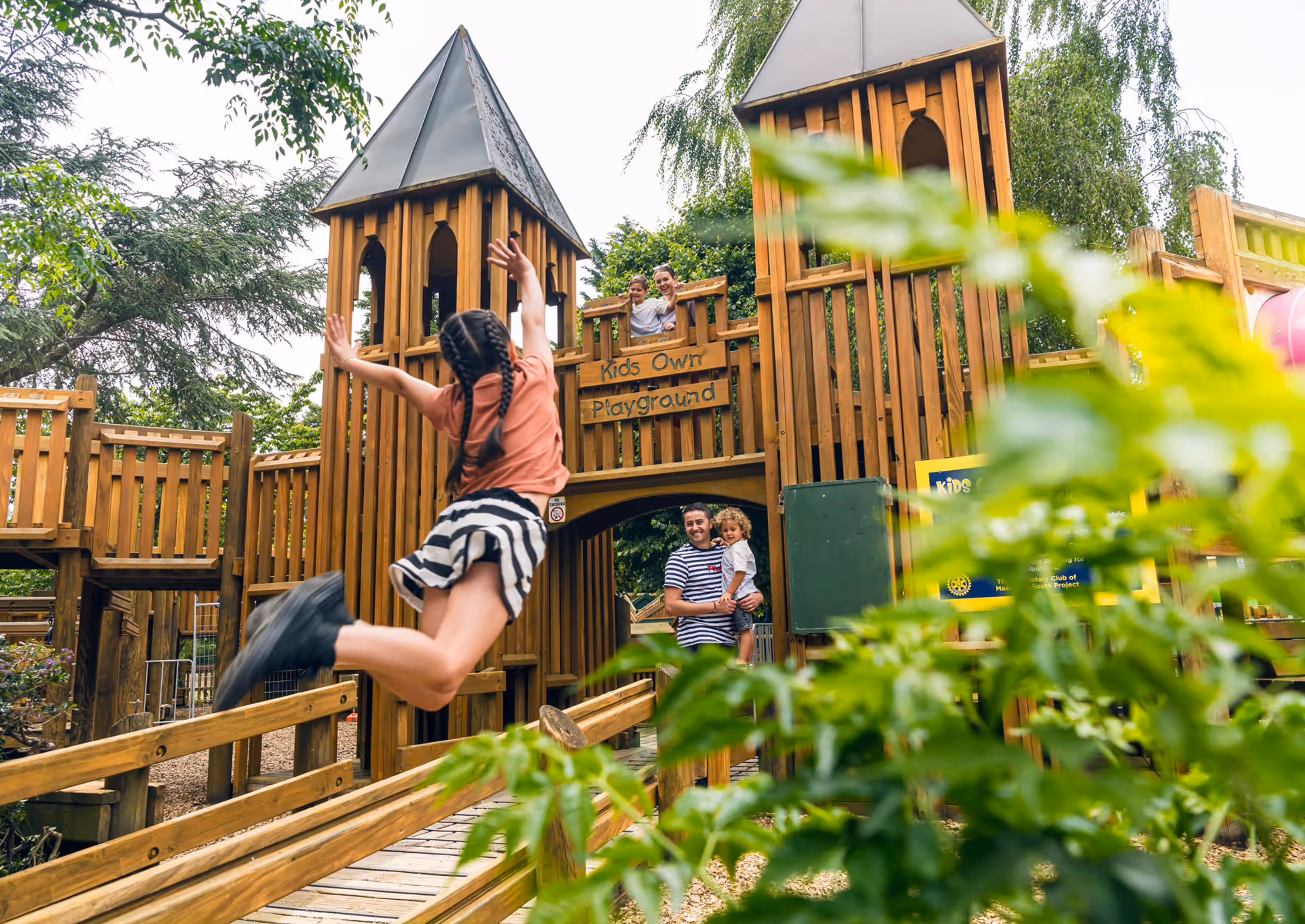 Child jumping with arms raised in front of a wooden playground structure labeled 'Kids Own Playground' with family members watching.