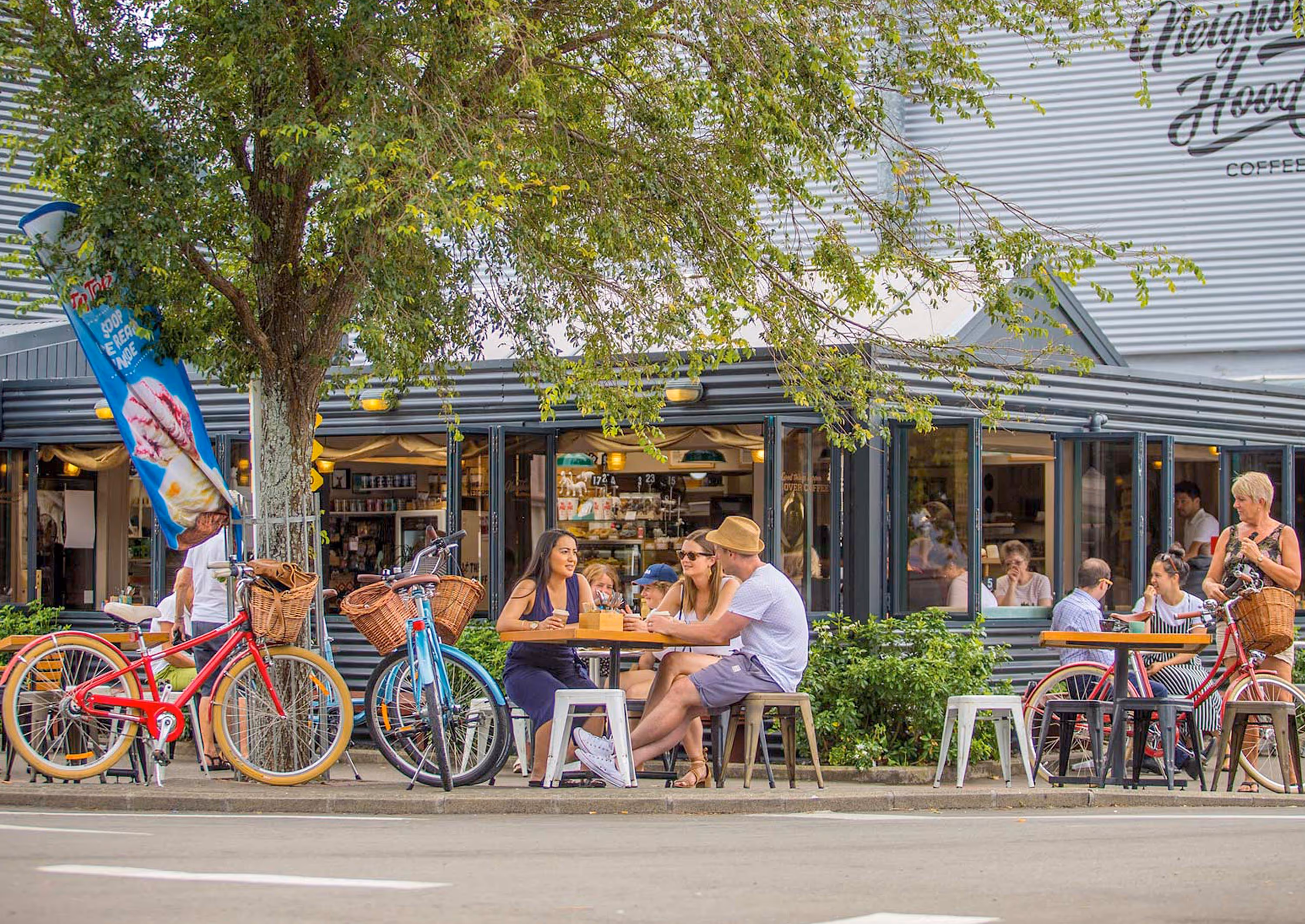 People sitting and standing outside a café with bicycles parked nearby under a leafy tree.