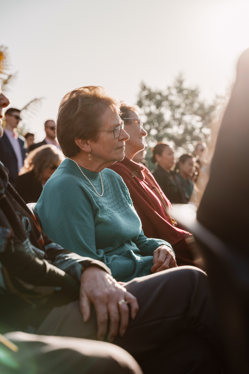 A wedding guest seated in the foreground watches the outdoor ceremony with quiet attention, warm sunlight glowing behind the crowd.