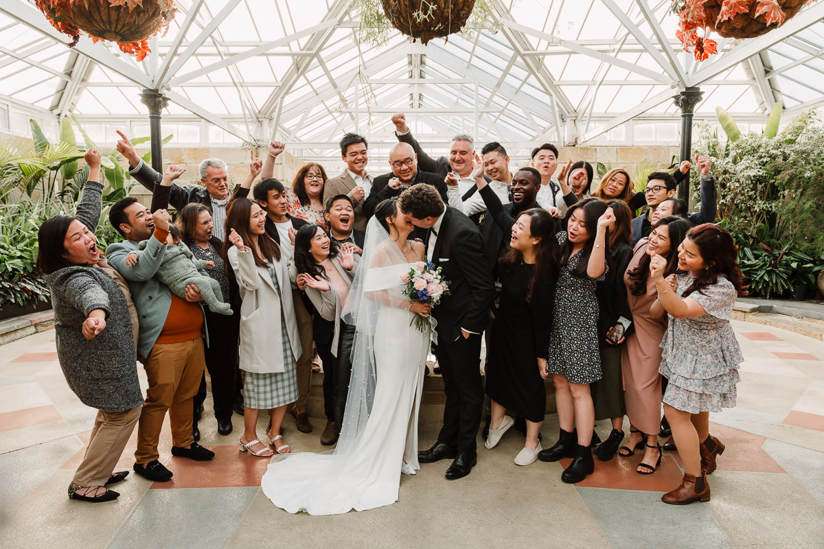 Bride and groom sharing a kiss in the centre of a cheering crowd of wedding guests inside the Royal Hobart Botanical Gardens conservatory, with the glass ceiling and tropical plants above.