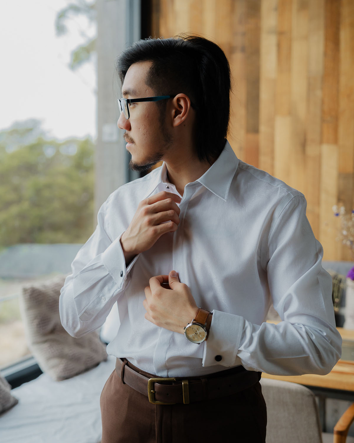 Man wearing glasses and a wristwatch buttoning up a white dress shirt indoors.
