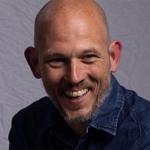 Peter Daxon smiling in a professional headshot, wearing a patterned dark shirt with a gray background.