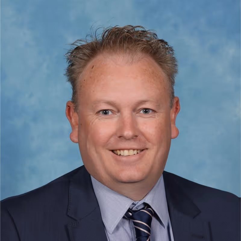 Professional headshot of Kieran Watson wearing a blue suit and striped tie with a light blue background.
