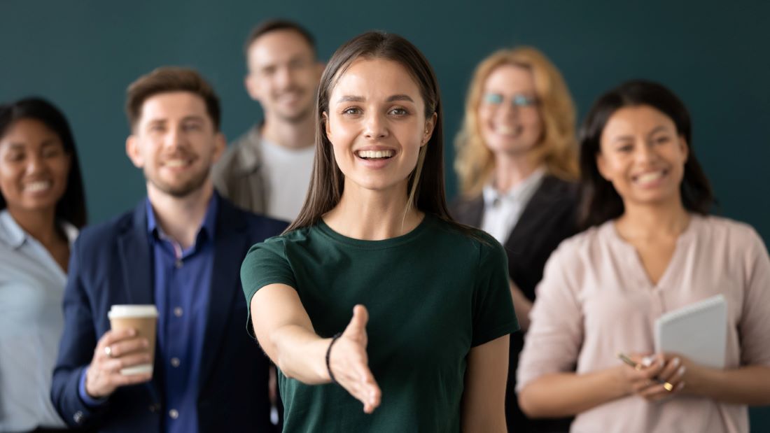 L’image montre un groupe de six personnes réunies dans un cadre professionnel et convivial. Au centre, une femme en chemise vert foncé tend la main vers l’avant, dans un geste d’accueil ou de poignée de main