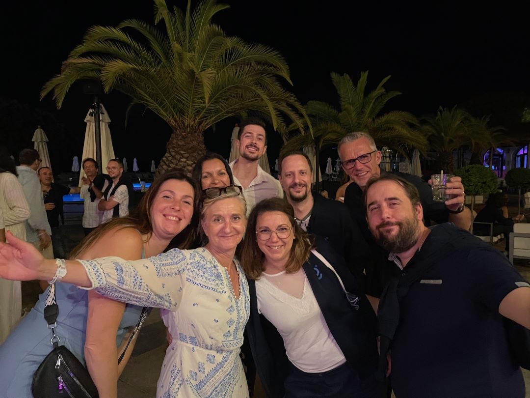 L’image montre un groupe de neuf adultes réunis pour une photo prise de nuit, dans un cadre extérieur évoquant une ambiance tropicale ou de resort