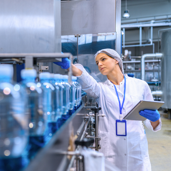 L’image montre un opérateur en blouse blanche, équipé d’un filet à cheveux, de gants bleus et d’un badge d’identification, en train d’inspecter une ligne de production automatisée